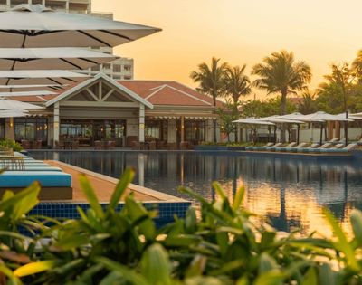 A resort swimming pool with rows of lounge chairs and umbrellas, overlooking a building with a tiled roof and palm trees, during sunset.