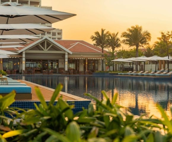 A resort swimming pool with rows of lounge chairs and umbrellas, overlooking a building with a tiled roof and palm trees, during sunset.