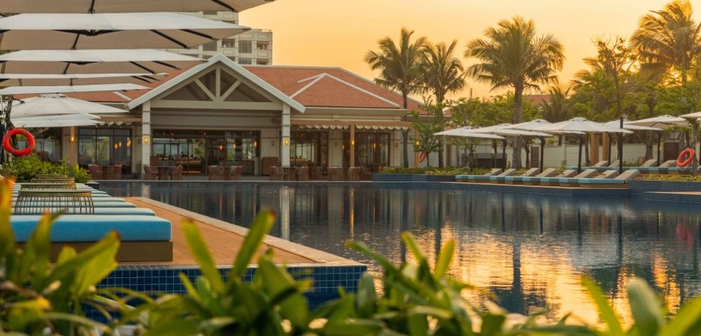 A resort swimming pool with rows of lounge chairs and umbrellas, overlooking a building with a tiled roof and palm trees, during sunset.