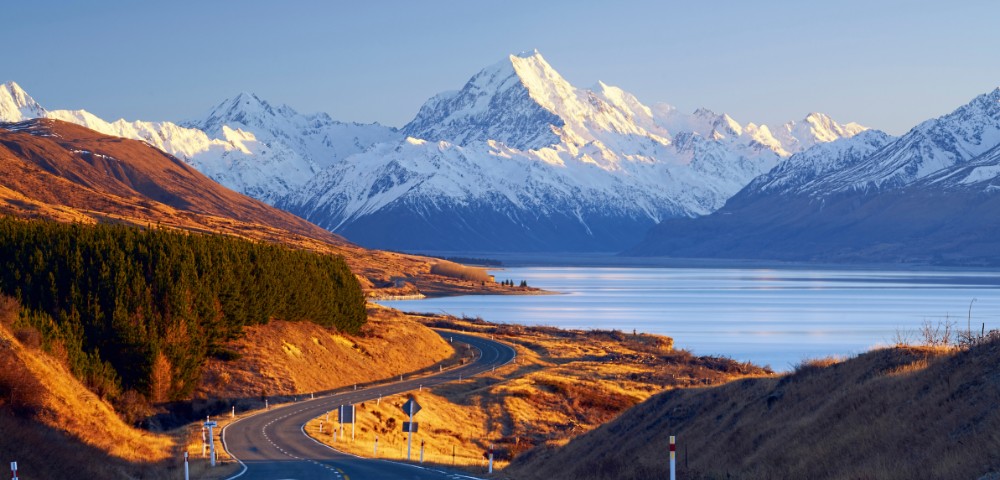 Winding road through golden grasslands leads to snowy mountains by a serene lake, under a clear blue sky at sunset. Majestic, peaceful landscape.