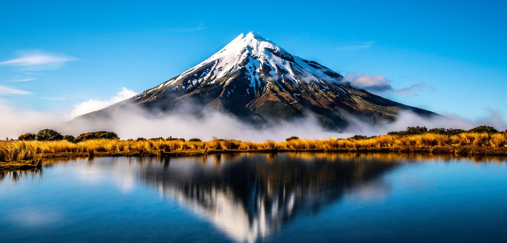 Snow-capped mountain reflected in a calm lake, surrounded by low clouds and golden reeds. The scene conveys serenity and natural beauty.