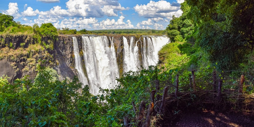 Majestic waterfall cascades over a cliff amid lush greenery, under a sky filled with fluffy clouds. The scene is vibrant and awe-inspiring.