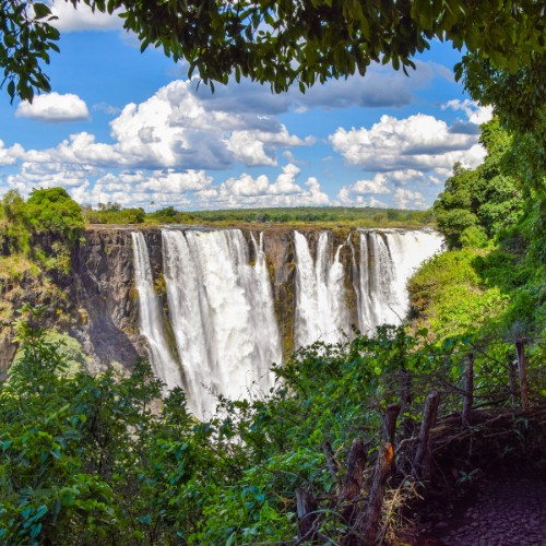 Majestic waterfall cascades over a cliff amid lush greenery, under a sky filled with fluffy clouds. The scene is vibrant and awe-inspiring.