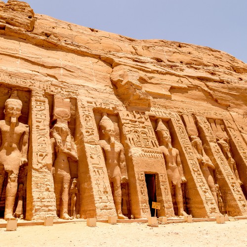 Historic Egyptian temple facade carved into a sunlit sandstone cliff, featuring large statues and detailed hieroglyphics under a clear blue sky.