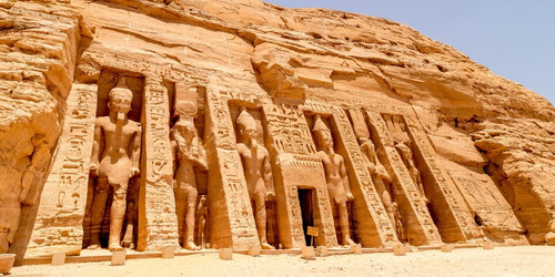 Historic Egyptian temple facade carved into a sunlit sandstone cliff, featuring large statues and detailed hieroglyphics under a clear blue sky.