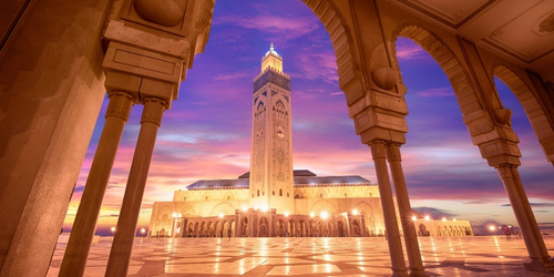 Sunset view of the Hassan II Mosque in Casablanca, framed by arches. The sky is a vibrant mix of purple, pink, and orange, creating a serene ambiance.