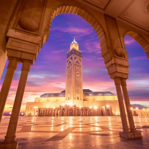 Sunset view of the Hassan II Mosque in Casablanca, framed by arches. The sky is a vibrant mix of purple, pink, and orange, creating a serene ambiance.