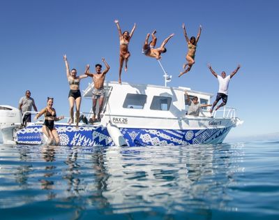 A group of people jump off the back of a white boat with blue patterned trim into clear blue water under a bright sky.