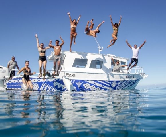 A group of people jump off the back of a white boat with blue patterned trim into clear blue water under a bright sky.