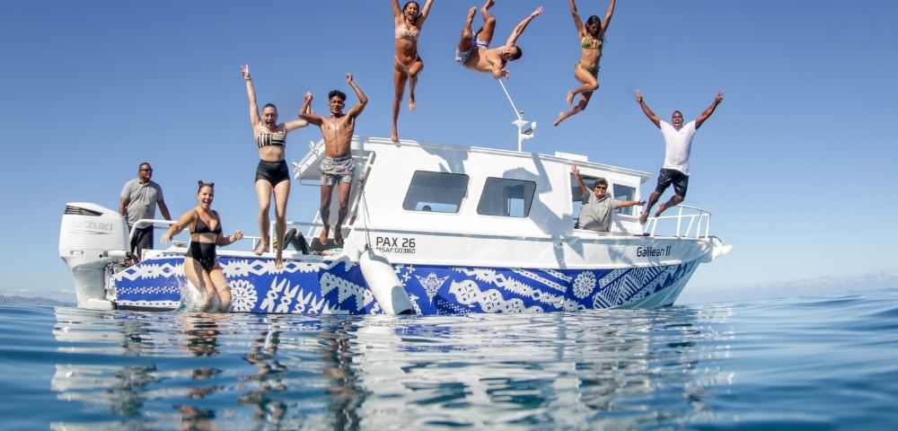 A group of people jump off the back of a white boat with blue patterned trim into clear blue water under a bright sky.