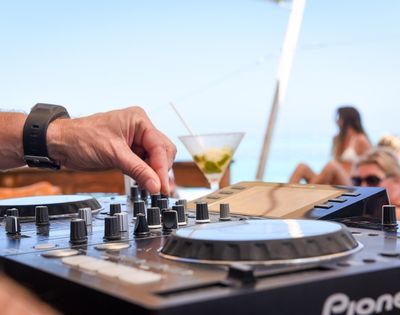 Close-up of a hand adjusting knobs on a DJ mixing board with a martini and blurred people in swimwear in the background, suggesting an outdoor party atmosphere.