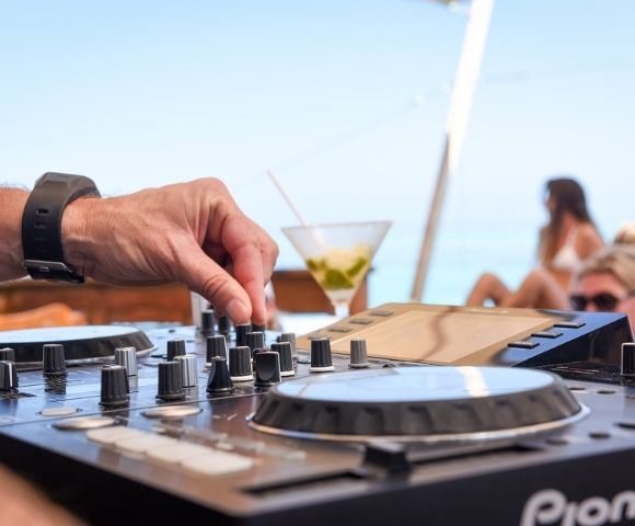 Close-up of a hand adjusting knobs on a DJ mixing board with a martini and blurred people in swimwear in the background, suggesting an outdoor party atmosphere.