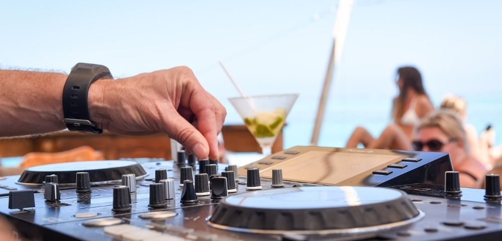 Close-up of a hand adjusting knobs on a DJ mixing board with a martini and blurred people in swimwear in the background, suggesting an outdoor party atmosphere.