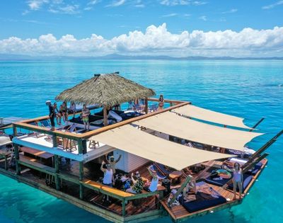 A multi-level floating wooden deck with thatched roof, sunshades, lounge chairs, and people enjoying the tropical ocean waters under a blue sky with scattered clouds.