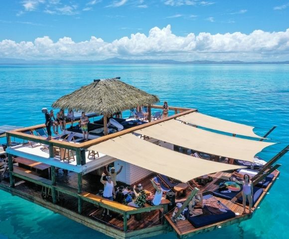 A multi-level floating wooden deck with thatched roof, sunshades, lounge chairs, and people enjoying the tropical ocean waters under a blue sky with scattered clouds.