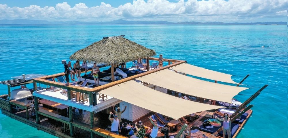 A multi-level floating wooden deck with thatched roof, sunshades, lounge chairs, and people enjoying the tropical ocean waters under a blue sky with scattered clouds.