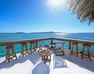 Oceanfront deck with wooden stools, lounge chairs, and tables under a thatched roof, overlooking clear blue water and distant islands.