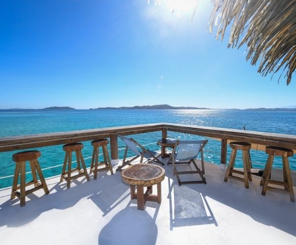 Oceanfront deck with wooden stools, lounge chairs, and tables under a thatched roof, overlooking clear blue water and distant islands.
