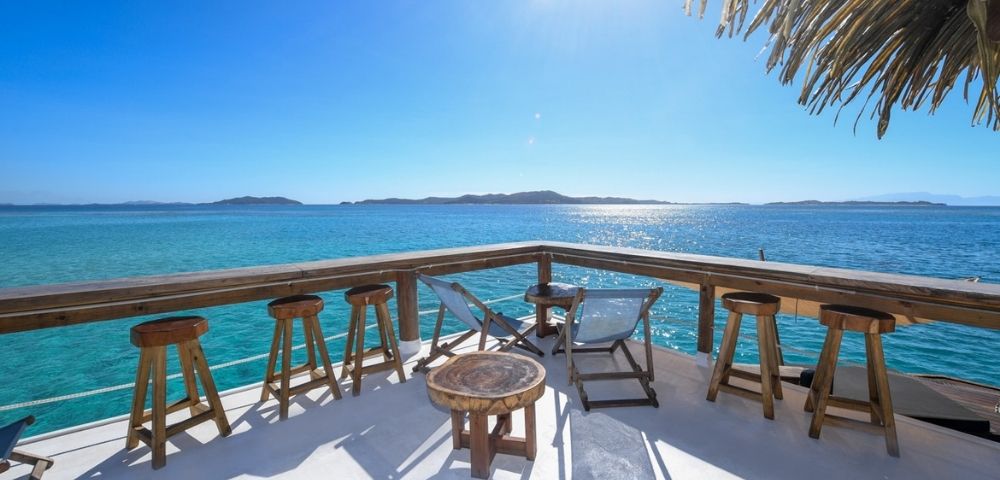 Oceanfront deck with wooden stools, lounge chairs, and tables under a thatched roof, overlooking clear blue water and distant islands.