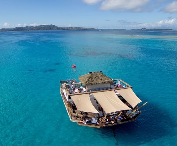 A floating wooden raft with shaded areas and people relaxing floats on clear turquoise ocean waters near distant islands.