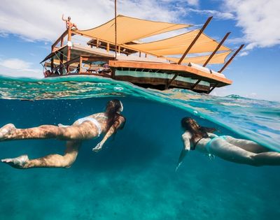 Two women swim underwater near a partially submerged floating deck with tan canopies. The scene conveys a sense of relaxation and adventure.