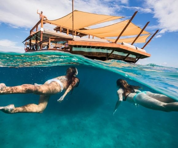 Two women swim underwater near a partially submerged floating deck with tan canopies. The scene conveys a sense of relaxation and adventure.