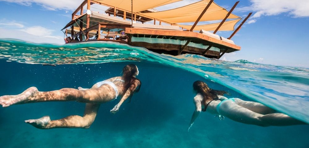 Two women swim underwater near a partially submerged floating deck with tan canopies. The scene conveys a sense of relaxation and adventure.