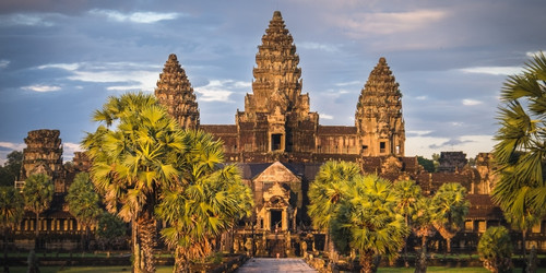 Ancient temple with intricate stone towers surrounded by lush palm trees and vibrant greenery under a partly cloudy sky, evoking a sense of grandeur.