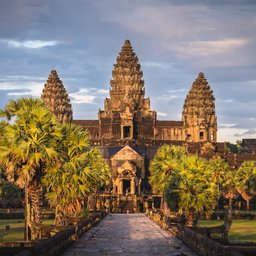 Ancient temple with intricate stone towers surrounded by lush palm trees and vibrant greenery under a partly cloudy sky, evoking a sense of grandeur.