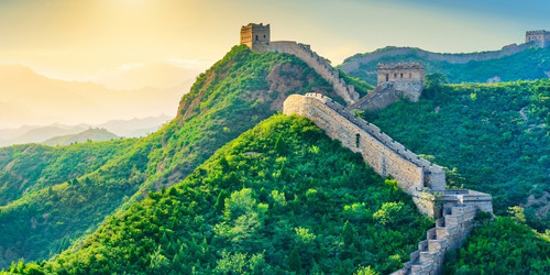 View of the Great Wall of China winding over lush green hills at sunrise, with a warm glow in the sky and layers of distant misty mountains.