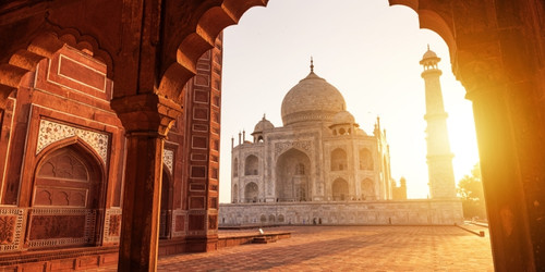 View of the Taj Mahal at sunrise through an ornate archway, with warm golden light illuminating the marble structure and casting long shadows.