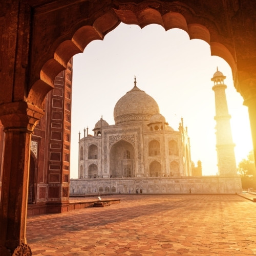 View of the Taj Mahal at sunrise through an ornate archway, with warm golden light illuminating the marble structure and casting long shadows.