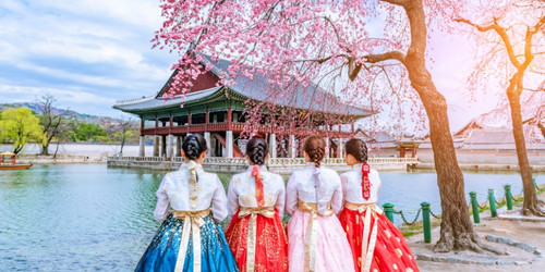 Four people in colorful hanboks stand by a serene pond, admiring a traditional Korean pavilion. Cherry blossoms and soft sunlight add a peaceful ambiance.
