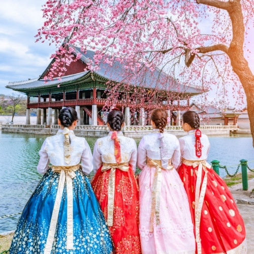 Four people in colorful hanboks stand by a serene pond, admiring a traditional Korean pavilion. Cherry blossoms and soft sunlight add a peaceful ambiance.