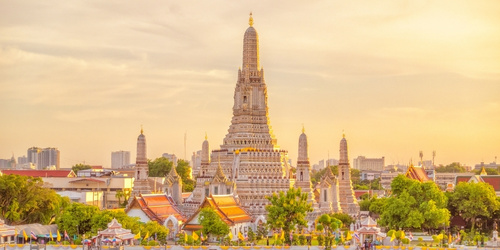 Sunset view of Wat Arun temple in Bangkok, highlighting its ornate spires and colorful rooftops. The river reflects the golden light, creating a serene atmosphere.