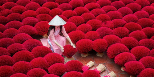 A woman in a pink dress and conical hat arranges vibrant red incense sticks laid out to dry on the ground. The scene is visually striking and peaceful.