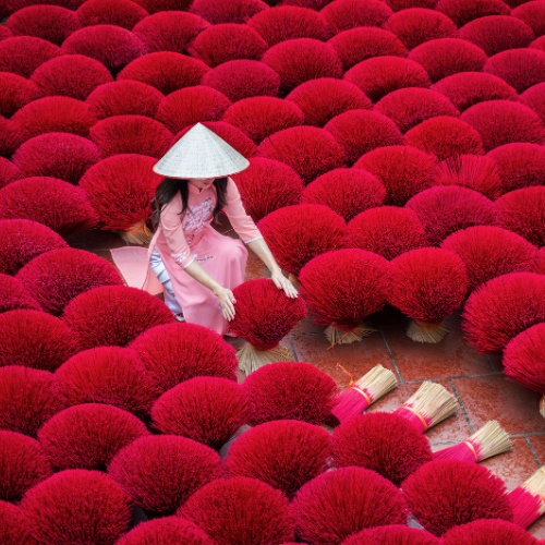 A woman in a pink dress and conical hat arranges vibrant red incense sticks laid out to dry on the ground. The scene is visually striking and peaceful.