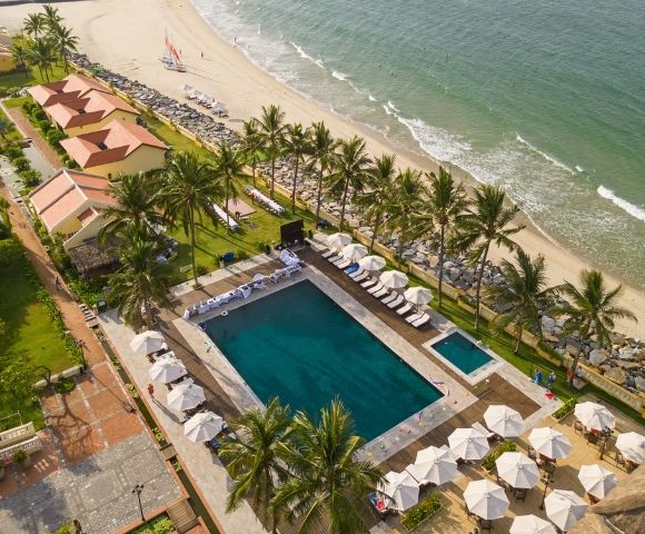 Aerial view of a beachfront resort with a large pool, palm trees, sun loungers, umbrellas, and sandy beach waves.