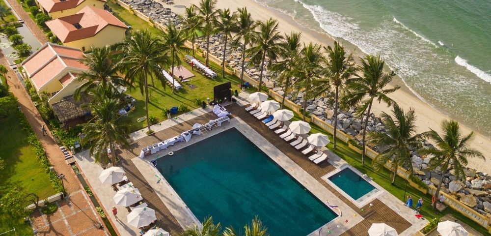 Aerial view of a beachfront resort with a large pool, palm trees, sun loungers, umbrellas, and sandy beach waves.