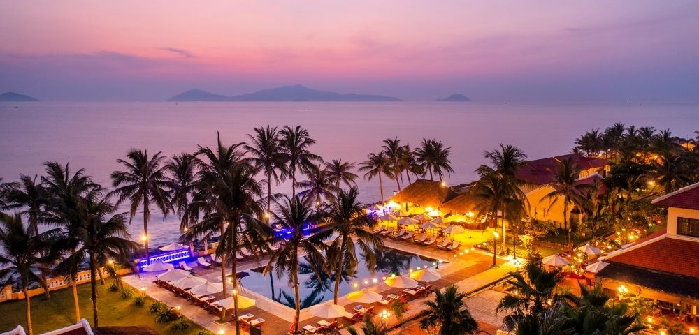 Tropical resort at dusk with illuminated pool, palm trees, beach, and calm sea under a pink and purple sky.