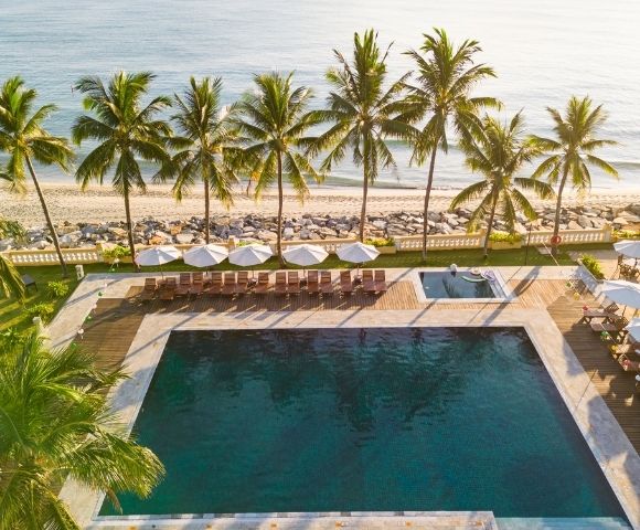 A serene beachfront pool area with lounge chairs, umbrellas, palm trees, and a hot tub overlooking a sandy beach and calm ocean.