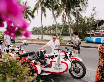 Two people interacting near vibrant flowers and a parked red and white motorcycle by a scenic waterfront in a tropical setting.