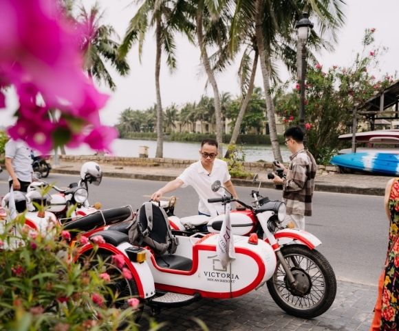 Two people interacting near vibrant flowers and a parked red and white motorcycle by a scenic waterfront in a tropical setting.