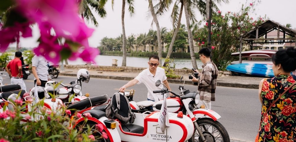 Two people interacting near vibrant flowers and a parked red and white motorcycle by a scenic waterfront in a tropical setting.