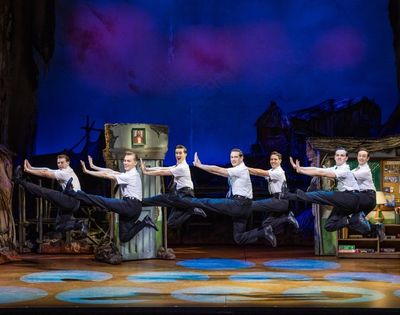 Seven male dancers in white shirts and black pants perform a synchronized high jump on a colorful stage with theatrical set pieces.