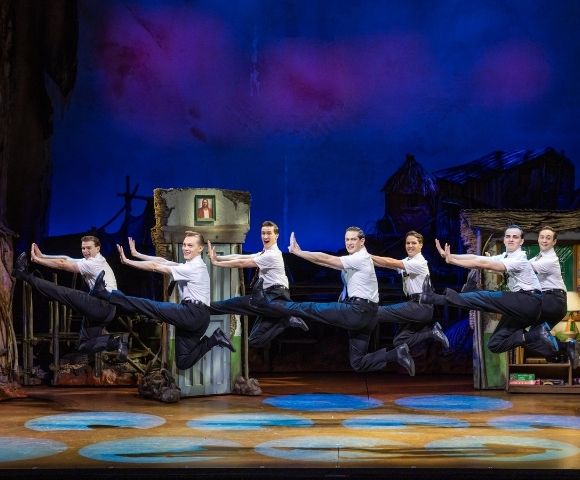 Seven male dancers in white shirts and black pants perform a synchronized high jump on a colorful stage with theatrical set pieces.