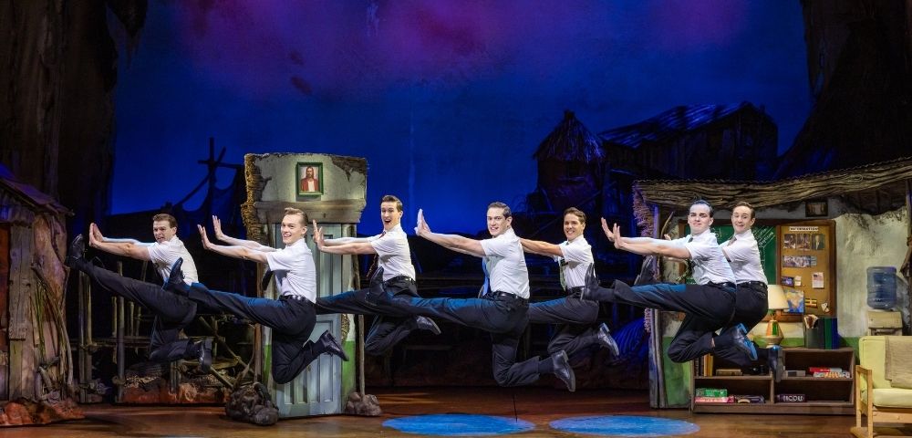 Seven male dancers in white shirts and black pants perform a synchronized high jump on a colorful stage with theatrical set pieces.