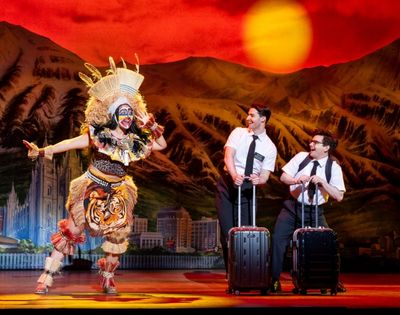 A performer in a colorful tribal costume dances energetically while two men in white shirts and ties stand with luggage against a vibrant sunset backdrop.