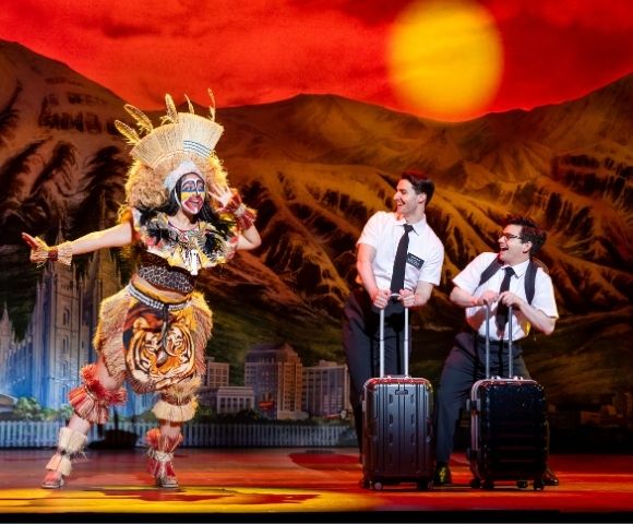 A performer in a colorful tribal costume dances energetically while two men in white shirts and ties stand with luggage against a vibrant sunset backdrop.