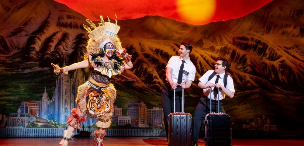 A performer in a colorful tribal costume dances energetically while two men in white shirts and ties stand with luggage against a vibrant sunset backdrop.